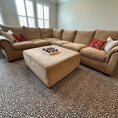 Sale Photo Thumbnail #26: Large beige fabric sectional sofa with nailhead trim, matching square ottoman, decorative throw pillows, and a patterned animal-print area rug.