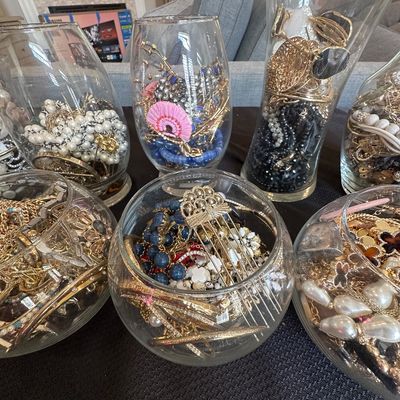 Sale Photo Thumbnail #4: Assorted costume jewelry arranged in clear glass vases and bowls, featuring gold-tone chains, beaded necklaces, bangles, and hair accessories.