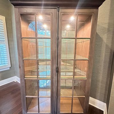 Sale Photo Thumbnail #114: Tall wooden display cabinet in a weathered grey-brown finish, featuring double glass-paneled doors, interior lighting, and adjustable glass shelves.