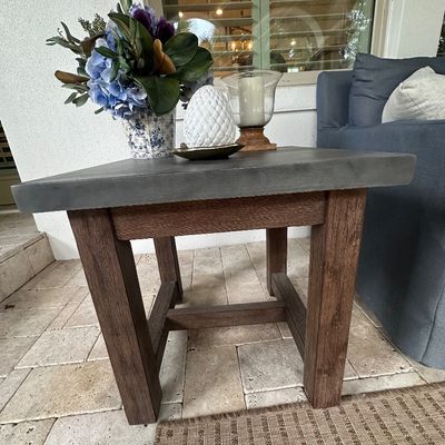 Sale Photo Thumbnail #22: Rustic modern patio end table featuring a thick concrete or stone top on a sturdy dark wood base. Decorative accents include a blue-and-white floral vase, a pinecone figurine, and a glass hurricane candle holder.