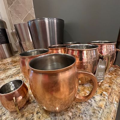 Sale Photo Thumbnail #41: Collection of copper Moscow Mule mugs, including smooth and hammered finishes, alongside stainless steel barware accessories.