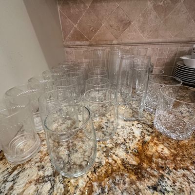 Sale Photo Thumbnail #39: Large collection of assorted clear glassware, including stemless wine glasses, highball glasses, and cut-crystal rocks glasses arranged on a granite countertop.