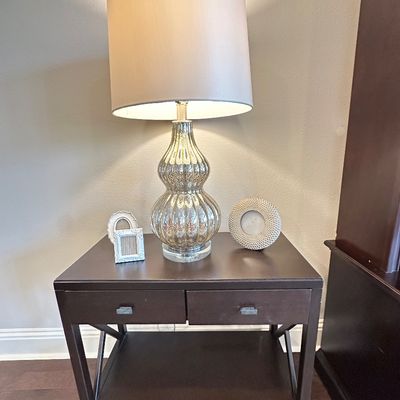 Sale Photo Thumbnail #62: Contemporary mercury glass table lamp with a neutral drum shade, resting on a dark wood end table alongside small decorative picture frames.