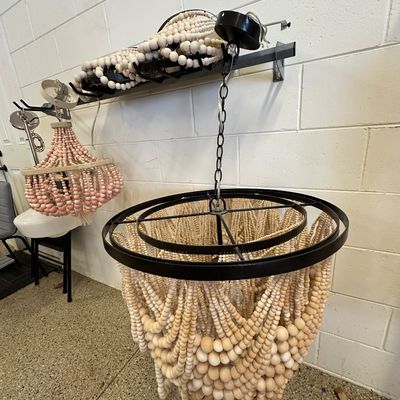 Sale Photo Thumbnail #122: Boho-style natural wooden bead pendant light fixtures suspended from wall racks, featuring a large tiered cream beaded chandelier in the foreground and a smaller pink and white version in the background.
