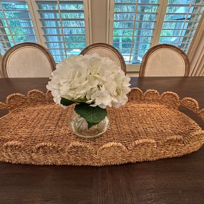 Sale Photo Thumbnail #48: Large woven seagrass tray with scalloped edges, holding a small clear glass vase of faux white hydrangeas on a dark wood dining table.