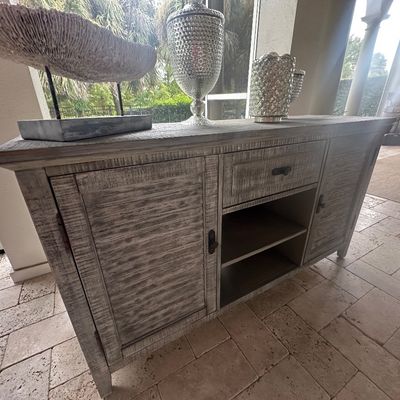 Sale Photo Thumbnail #70: Rustic gray-washed wood sideboard with panel doors, a center drawer, and open shelving, topped with decorative home accents.