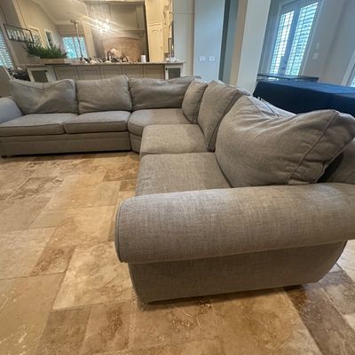 Sale Photo Thumbnail #91: Large contemporary light gray L-shaped sectional sofa with rolled arms and plush back cushions, set on a travertine tile floor.