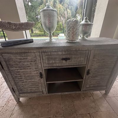 Sale Photo Thumbnail #69: Rustic weathered gray wooden sideboard cabinet, featuring two side doors, a central drawer, open shelving, and decorated with silver decorative urns and coral sculpture.