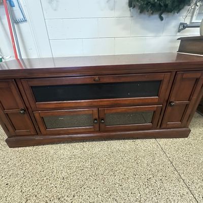 Sale Photo Thumbnail #94: Dark cherry wood media console featuring glass-paneled cabinet doors, a pull-down media compartment, and side storage doors.