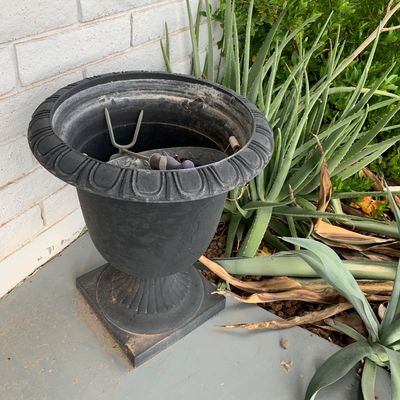 Sale Photo Thumbnail #133: Black urn-style outdoor planter with a square pedestal base and egg-and-dart trim detail, shown outdoors near garden foliage.