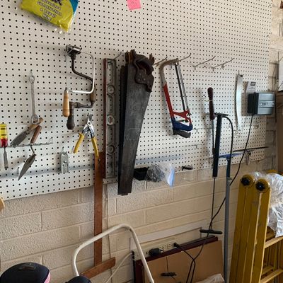 Sale Photo Thumbnail #56: Assorted vintage hand tools hanging on a pegboard wall, including handsaws, a coping saw, a hand drill, a level, and pliers, with folding ladders nearby.
