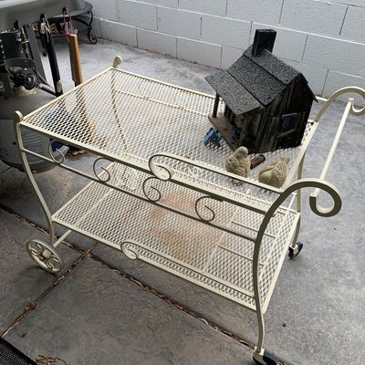 Sale Photo Thumbnail #36: Vintage white painted wrought iron patio tea cart with mesh shelves and scrollwork accents, holding a decorative wooden birdhouse and small bird figurines.