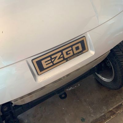 Sale Photo Thumbnail #46: Front view of a white EZGO golf cart featuring a prominent gold-and-black logo plate.