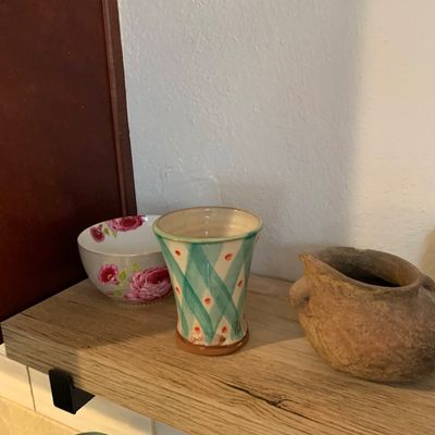 Sale Photo Thumbnail #76: Collection of decorative pottery pieces displayed on a wooden shelf, including a hand-painted floral bowl, a teal criss-cross pattern cup, and a rustic earthy clay vessel.