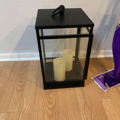 Sale Photo Thumbnail #58: Black metal lantern with clear glass panes, featuring a top loop handle and containing two cream-colored flameless pillar candles.