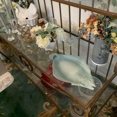 Sale Photo Thumbnail #81: A brass and glass tiered console table displaying various decorative items, including floral arrangements, candlesticks, and a light blue ceramic fish-shaped platter.