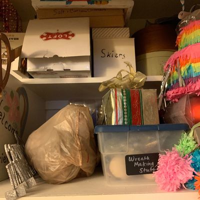 Sale Photo Thumbnail #100: Assorted holiday decor and craft supplies stacked on white closet shelves, including a plastic bin labeled "Wreath Making Stuff," gift boxes, and colorful paper decorations.