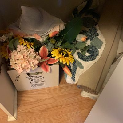 Sale Photo Thumbnail #105: Cardboard box filled with assorted artificial flowers alongside rolled patterned accent rugs stored under a shelf.