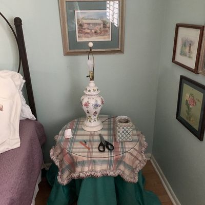 Sale Photo Thumbnail #85: Traditional floral painted ceramic urn-style table lamp resting on a skirted round nightstand, shown with framed artwork on the surrounding walls.
