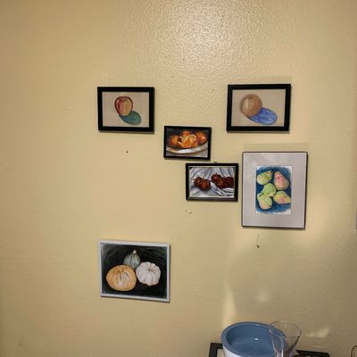 Sale Photo Thumbnail #29: Collection of framed original still life fruit artwork displayed on a yellow wall, accompanied by glassware on a table below.