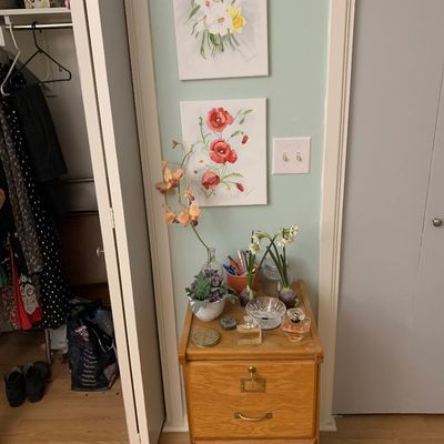 Sale Photo Thumbnail #111: Two-drawer oak filing cabinet topped with perfume bottles, artificial floral arrangements, and trinkets, alongside two wrapped canvas floral art prints on the wall.