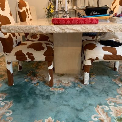 Sale Photo Thumbnail #14: Large travertine dining table with a thick rough-edge slab top, paired with dining chairs in brown and white cowhide fabric covers, set over a teal floral area rug.