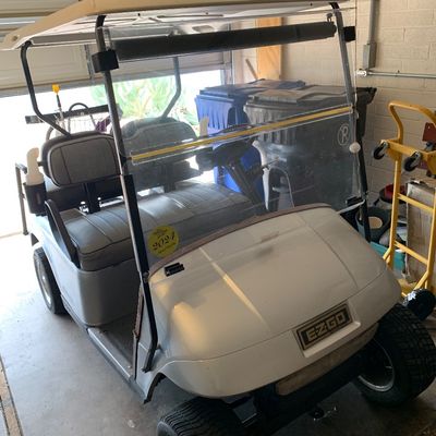 Sale Photo Thumbnail #45: EZGO golf cart featuring a light gray body, dual-row bench seating, overhead canopy, and folding windshield, parked inside a garage.