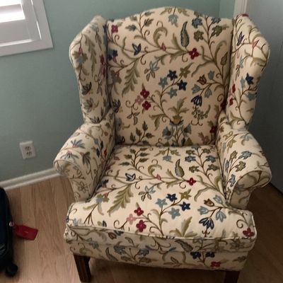 Sale Photo Thumbnail #84: Upholstered wingback armchair featuring a floral and vine crewelwork-style pattern on a cream background, supported by dark wooden legs.