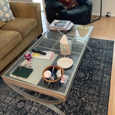 Sale Photo Thumbnail #2: Contemporary glass-top coffee table with a metallic frame, set atop a distressed dark patterned area rug in a living room.