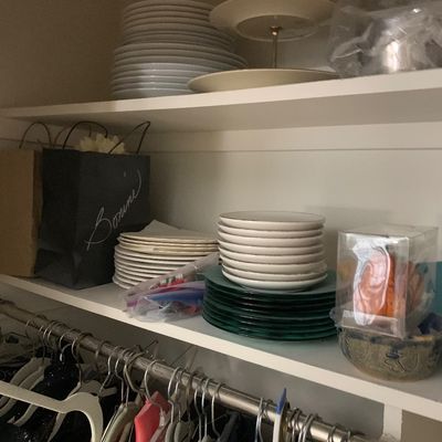 Sale Photo Thumbnail #112: Closet shelves filled with various stacks of ceramic and glass plates, a two-tiered brass serving tray, and assorted items above a hanging clothes rack.