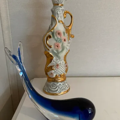 Sale Photo Thumbnail #16: Blue and clear art glass whale figurine alongside an ornate porcelain decanter with gold gilt scrollwork and painted floral details.