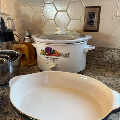 White ceramic oval baking dish set on a granite countertop, with a vintage Proctor-Silex slow cooker displaying a vegetable motif in the...