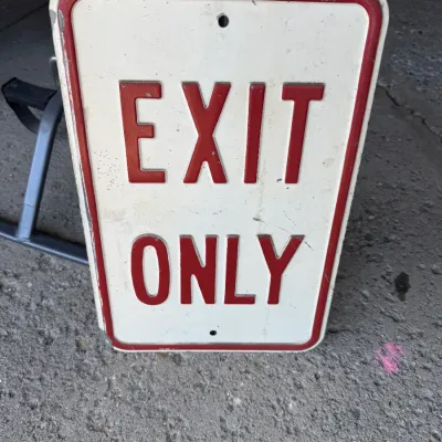 Vintage metal sign reading "EXIT ONLY" with embossed red lettering and a matching red border on a white background, showing...