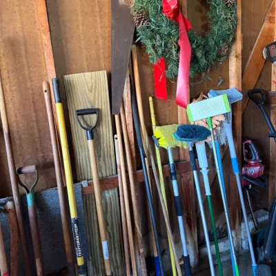 Collection of long-handled yard and cleaning tools including shovels, brooms, squeegees, and a hand saw, stored in a shed beside a...