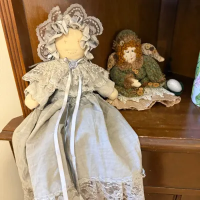 Two handmade folk art rag dolls sitting on a wooden shelf, including a larger doll in a grey bonnet and lace-trimmed dress alongside a...