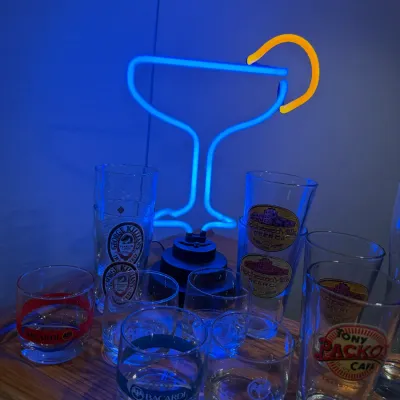 Sale Photo Thumbnail #20: A light-up neon margarita glass desk lamp illuminated in blue and yellow, displayed alongside various branded beer and liquor glassware.