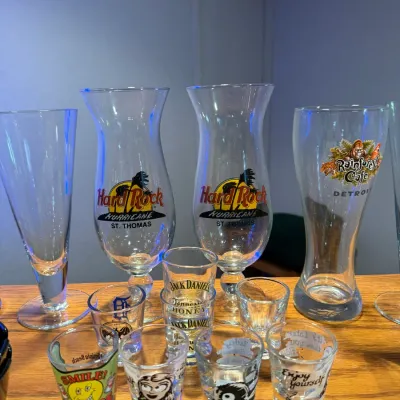 Sale Photo Thumbnail #21: Collection of souvenir glassware, including Hard Rock Cafe St. Thomas hurricane glasses, a Rainforest Cafe Detroit glass, and various novelty shot glasses.