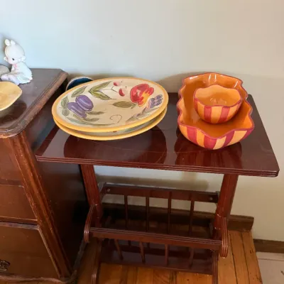 Vintage dark wood magazine rack end table displaying hand-painted fruit-patterned serving platters and orange-and-red striped ceramic bowls.
