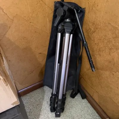 Collapsible aluminum camera tripod resting against a corner wall next to its black fabric carrying bag.