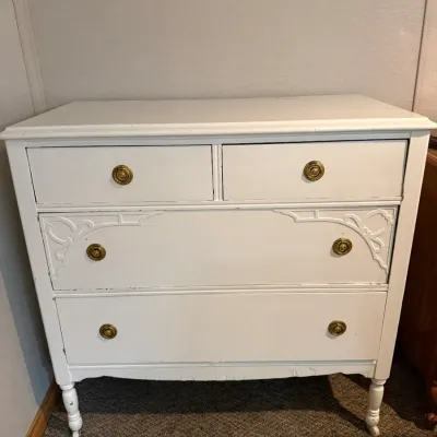Sale Photo Thumbnail #10: Vintage white painted wooden dresser featuring four drawers, ornate brass hardware, decorative carved corner details, and turned legs with casters.