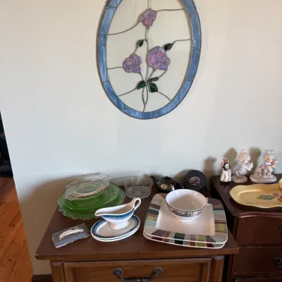 Oval stained glass suncatcher with a floral motif hanging above a wooden nightstand with assorted glassware and ceramics.