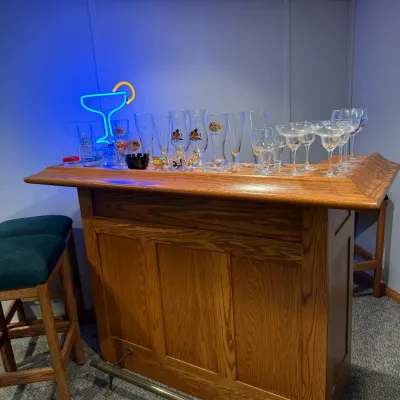 Sale Photo Thumbnail #19: Oak home bar unit with a brass footrail, accompanied by green upholstered barstools, a neon martini sign, and assorted glassware.