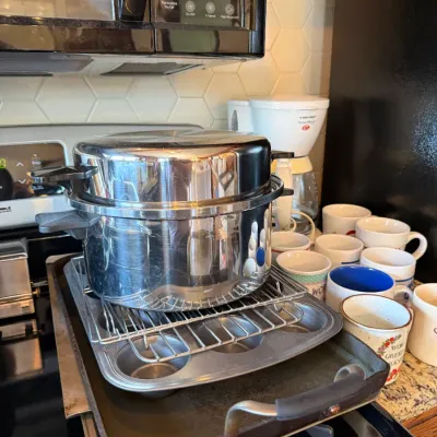 Large stainless steel stockpot with steamer insert resting on a cooling rack and muffin tin, surrounded by an assortment of ceramic...