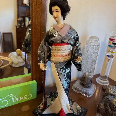 Vintage Japanese geisha doll dressed in a patterned black and silver kimono, mounted on a black base.