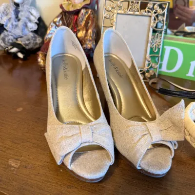 Pair of Kelly & Katie champagne-colored fabric peep-toe high heels featuring bow accents on the toes, presented on a wooden dresser...