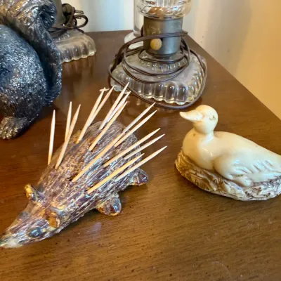 Novelty ceramic porcupine toothpick holder shown holding toothpicks, alongside a small carved duck figurine.