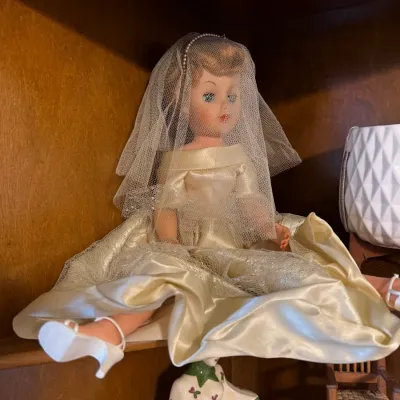 Vintage bride fashion doll in an off-white satin and tulle gown with matching veil and white heels, seated on a wooden display shelf.