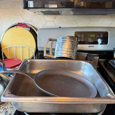 Collection of metal bakeware and cookware, including a cast iron griddle, stainless steel chafing pan, metal sifter, and loaf pan resting...
