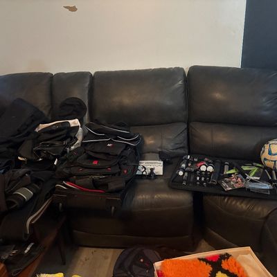 Sale Photo Thumbnail #8: Large black leather sectional couch filled with various household items, including luggage, backpacks, small hand tools, and a soccer ball.
