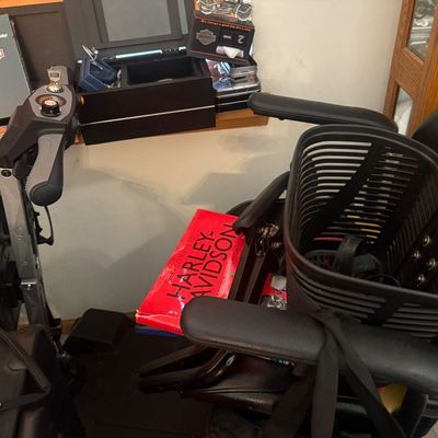 Sale Photo Thumbnail #27: Black mobility scooter with a front basket, padded seat, handlebars with controls, and a red Harley-Davidson history book placed on the floorboard.